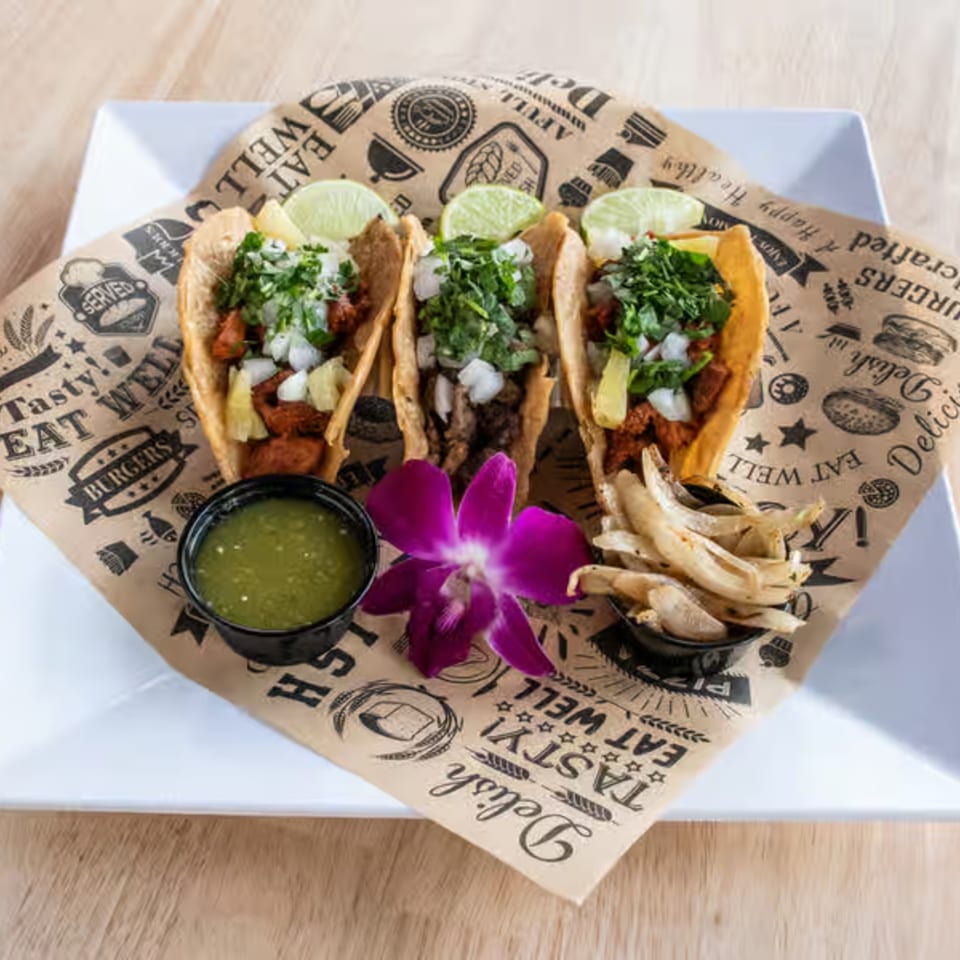 Three Maggy's House tacos with lime, cilantro, onions, green sauce, and grilled onions on a branded paper.