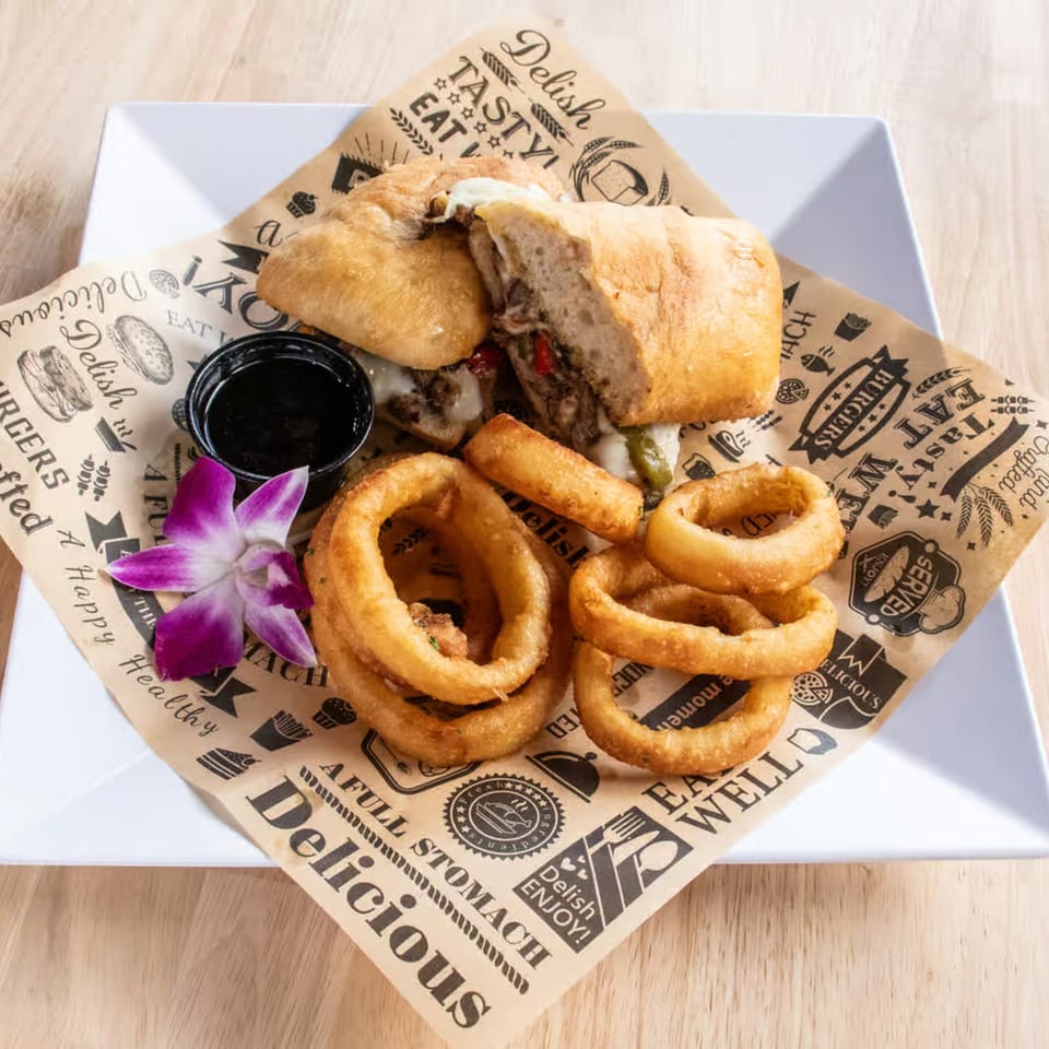 A Maggy's House steak and cheese sub with onion rings and a purple orchid on a white plate.
