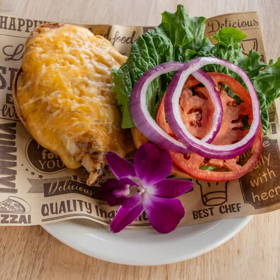 Grilled chicken breast topped with melted cheese, lettuce, tomato, and red onion, garnished with an orchid.